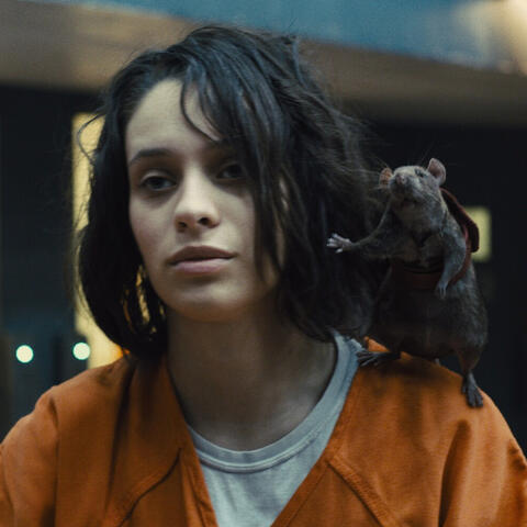 A photograph of a woman. She has dirty dark hair and an orange prisoners outfit. There is a cgi rat on her shoulder. The rat has a backpack.